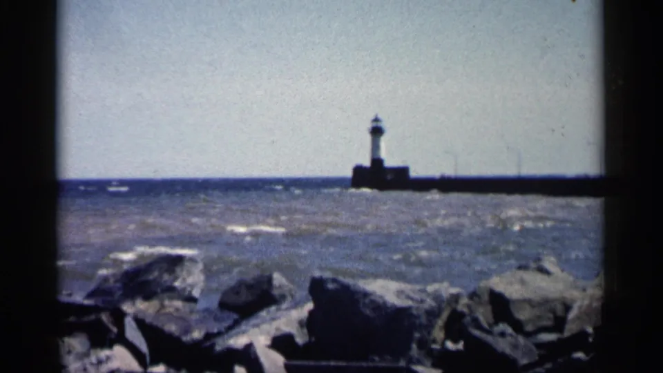 1961 Minnesota 8mm frame showing a lighthouse at the end of a pier across choppy water, with dark rocks in the foreground.
