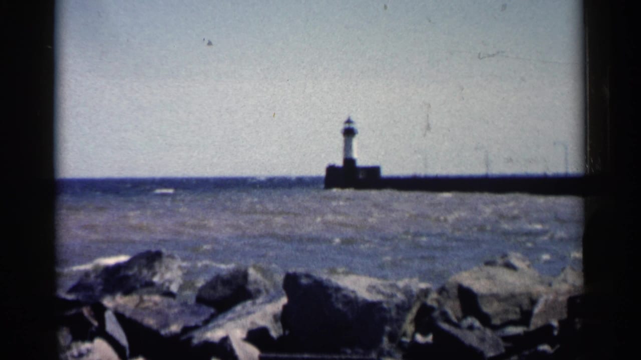 Lighthouse and pier across open water with large rocks in the foreground.