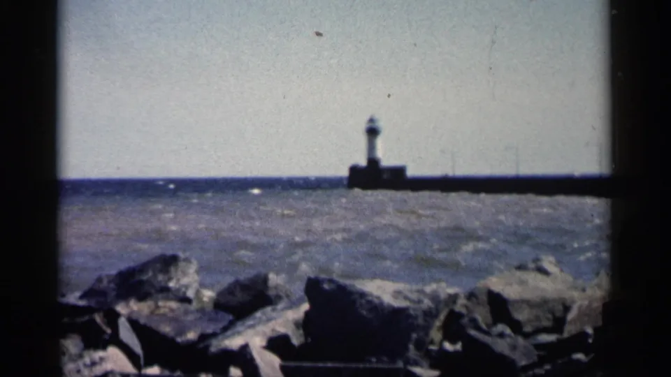 1961 Minnesota home-movie frame with shoreline rocks in front and a small lighthouse set on a long pier across the water.