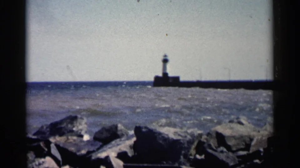 1961 Minnesota 8mm shot of a lighthouse and breakwater across choppy water, with dark shoreline rocks at the bottom edge.