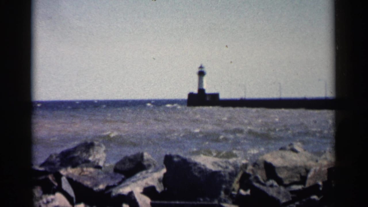 Lighthouse at the end of a pier beyond rocks and choppy water.
