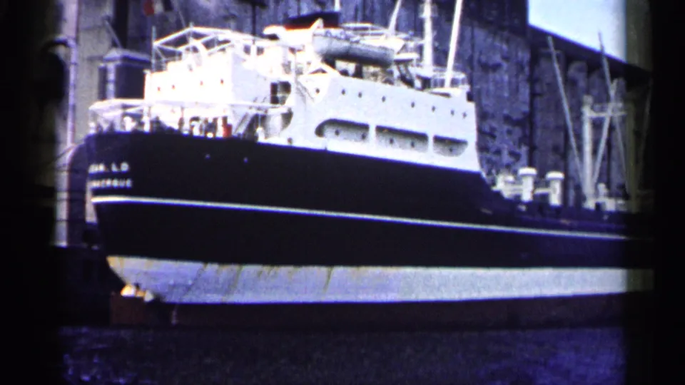 1961 Minnesota 8mm frame showing a large docked ship with a dark hull, white superstructure, railings, and a stone industrial wall behind it.