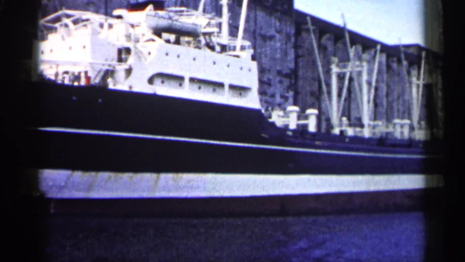 1961 Minnesota 8mm frame showing the ship's long dark hull, white upper deck area, and industrial wall as the camera pans along the vessel.
