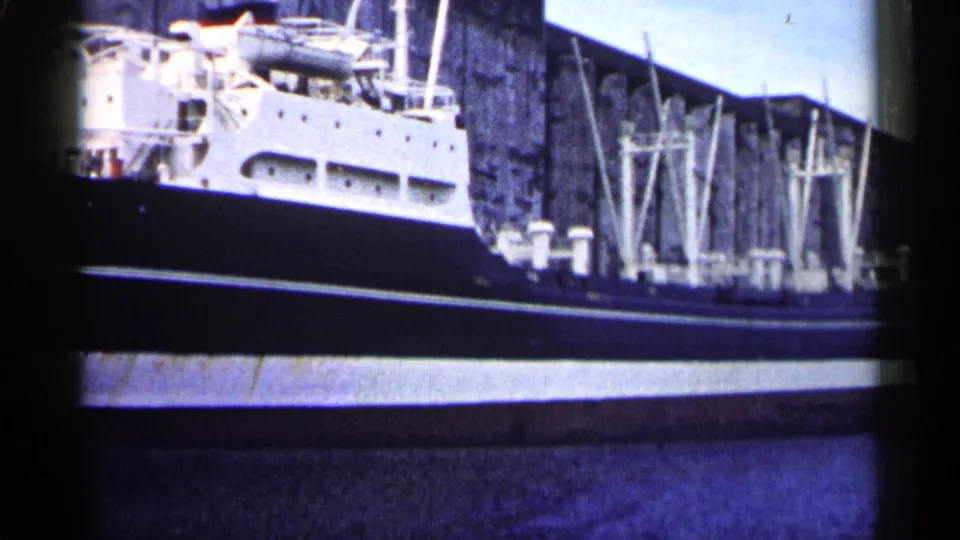 1961 Minnesota 8mm frame of a large moored vessel with railings, portholes, dockside structures, and a stone industrial wall in view.