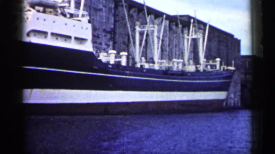 Blue-tinted 1961 Minnesota 8mm frame with a docked cargo ship, dark hull, white upper deck, rigging, pier edge, and water.