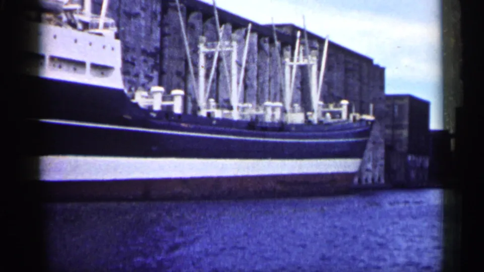 Final 1961 Minnesota 8mm frame showing the cargo ship still moored beside industrial dock structures with blue water below.