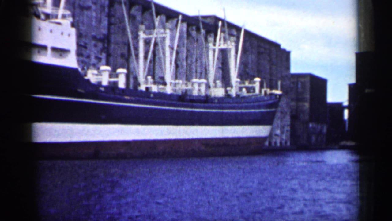 1961 Minnesota 8mm frame showing a large cargo ship docked beside industrial waterfront structures and still water.
