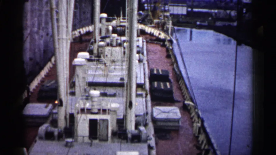 High-angle 1961 Minnesota view of a docked cargo ship deck, with mast hardware, railings, covered cargo, and water alongside.