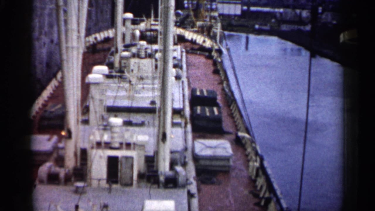 Cargo ship deck beside a narrow harbor channel with tall mast or crane structures.