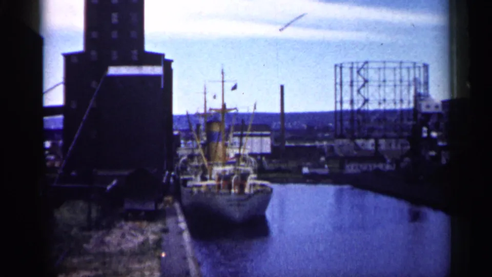 1961 Minnesota 8mm footage shows a docked vessel in an industrial harbor with water, a dark building mass, and a gas holder in the background.