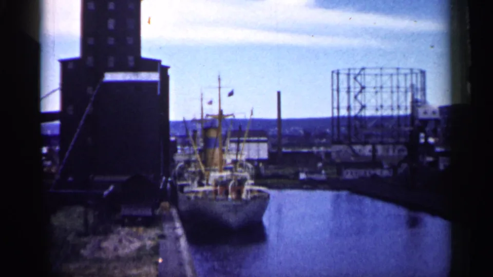 A faded 1961 Minnesota harbor frame shows a small docked vessel, industrial waterfront structures, open water, and a distant gas holder.
