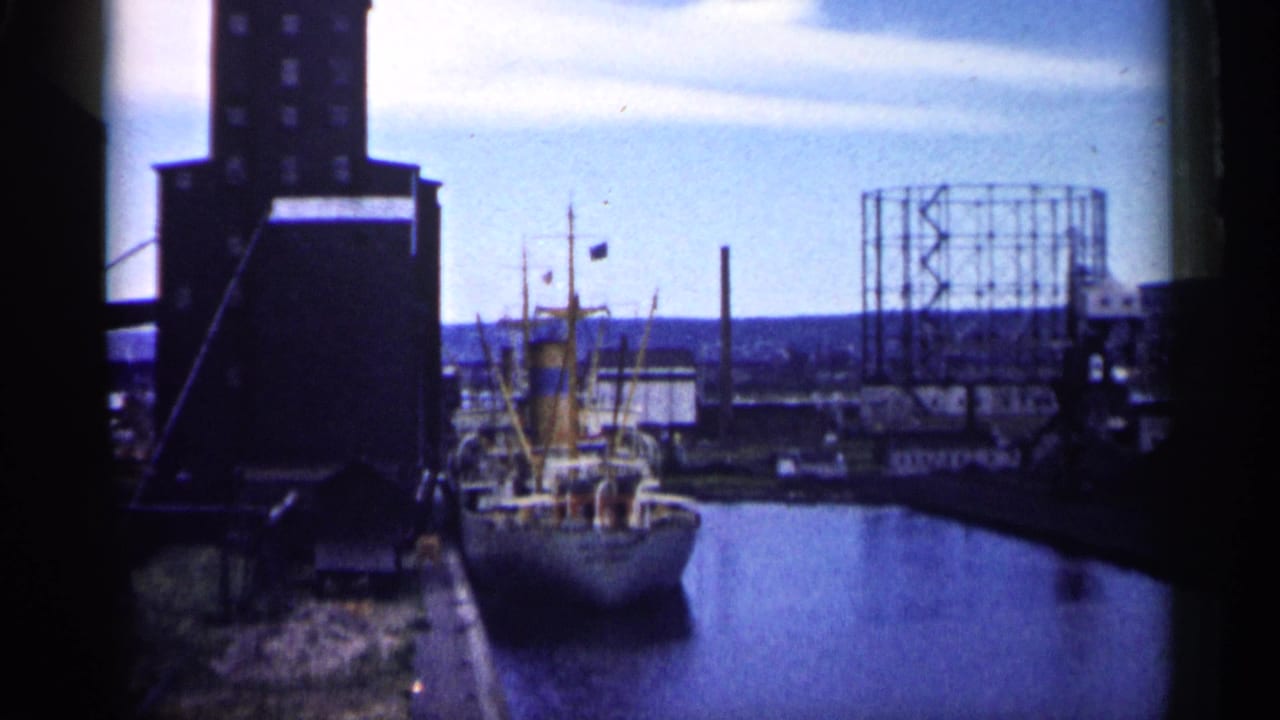 Freighter docked in an industrial harbor near a tall building and gas holder frame.