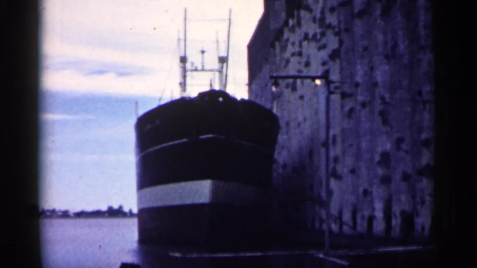Close harbor view of a dark-hulled freighter beside a tall masonry dock wall in 1961 Minnesota footage.