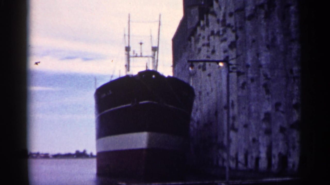 1961 8mm frame showing a dark-hulled ship with a white stripe docked next to a weathered stone pier, likely in Minnesota.