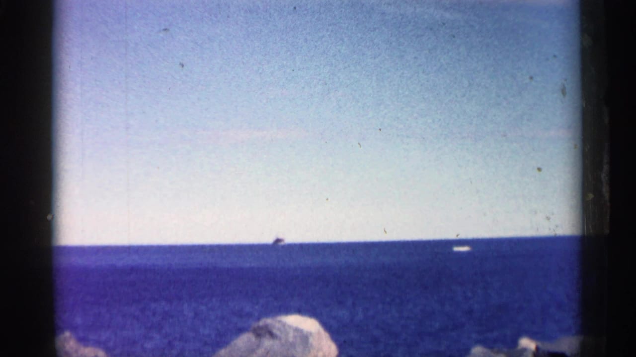 Wide 8mm view of blue open water, a distant vessel near the horizon, and pale shoreline rocks in the foreground.