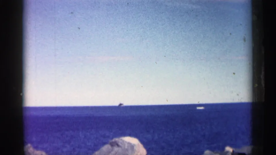 Wide 8mm view of blue open water, a distant vessel near the horizon, and pale shoreline rocks in the foreground.