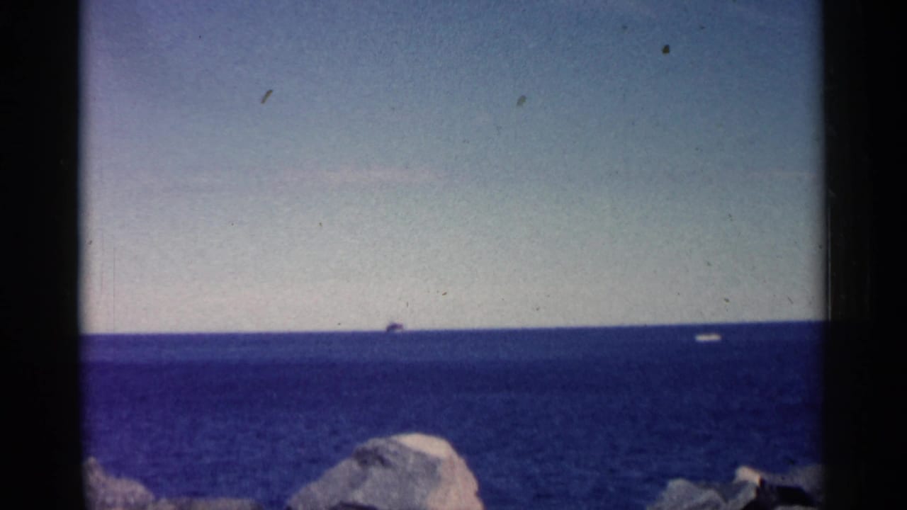 Blue water horizon with a small distant ship and shoreline rocks at the bottom edge.