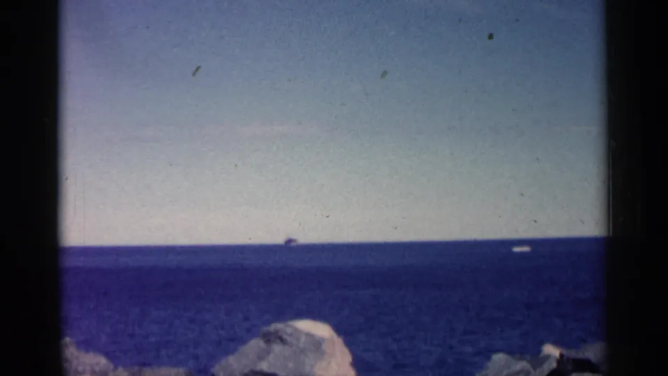 1961 Minnesota 8mm frame showing a small vessel far out on blue water, with pale sky and rocky shoreline in the foreground.