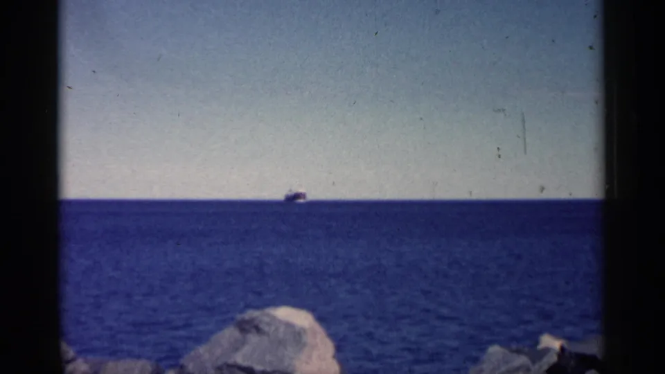 1961 Minnesota home-movie frame with a distant ship silhouetted near the horizon over calm blue water and low rocks.