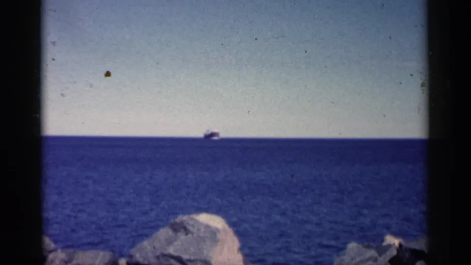 1961 Minnesota 8mm frame showing a wide blue water surface, a tiny ship at the horizon, and rocky shoreline at the bottom.