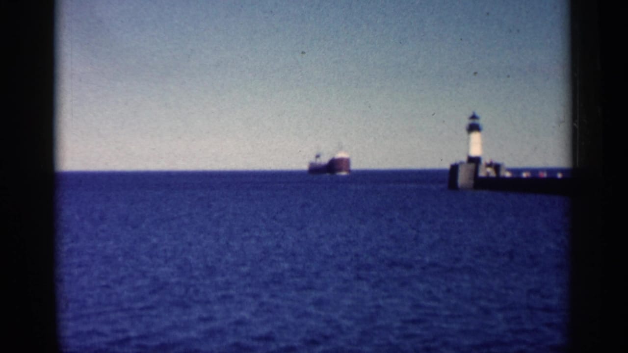 Distant freighter on blue water near a lighthouse on a pier.