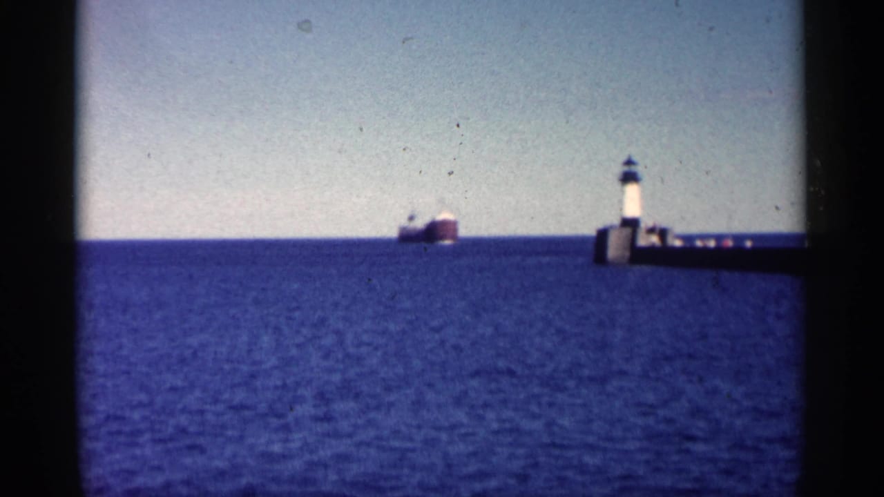 Wide 8mm view of a freighter offshore with a lighthouse and breakwater visible on the right in Minnesota footage from 1961.