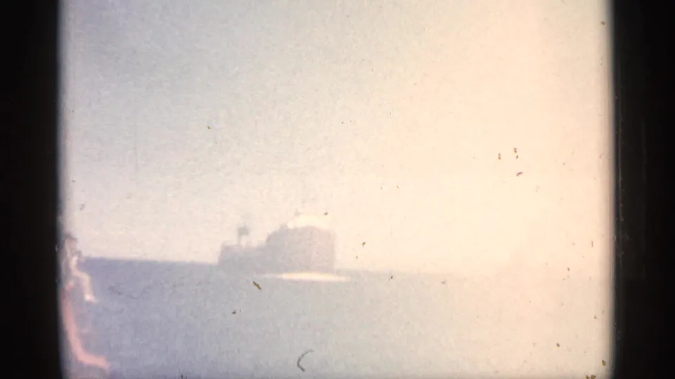 Bright, washed 8mm frame with the freighter faintly visible across the water and shoreline or railing detail along the left edge.