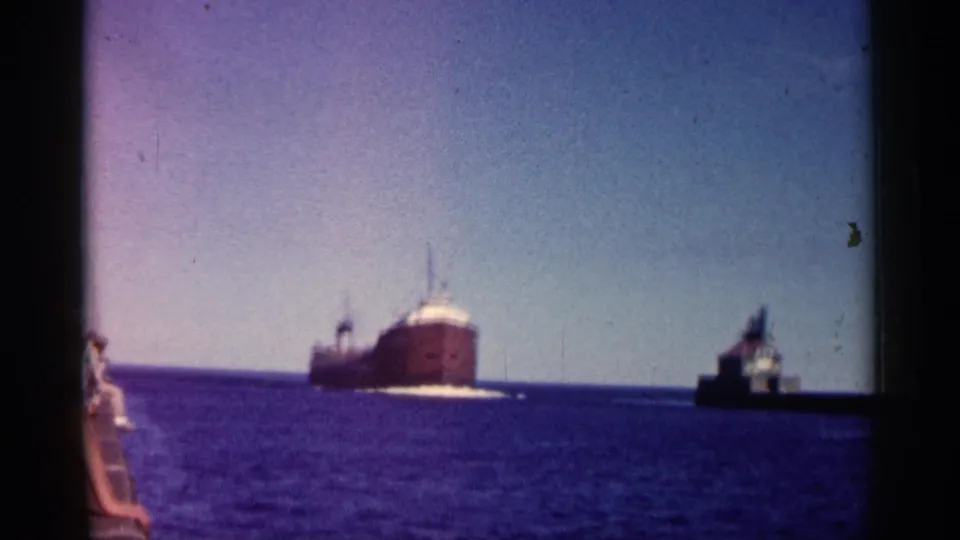 Closer 1961 Minnesota 8mm view of a large freighter moving past a lighthouse and pier, with blue water and film gate edges visible.