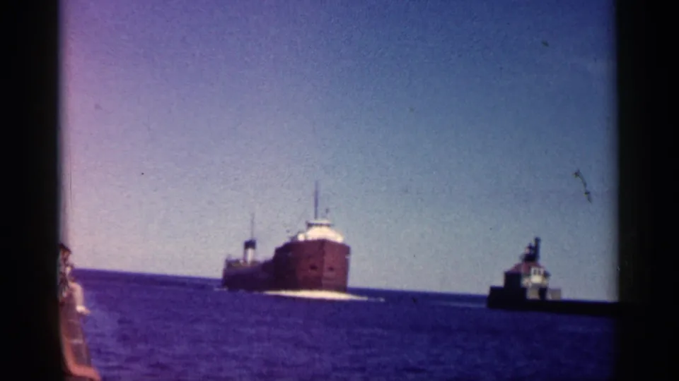 1961 Minnesota 8mm still showing a large freighter passing on calm water, with a small harbor structure nearby.