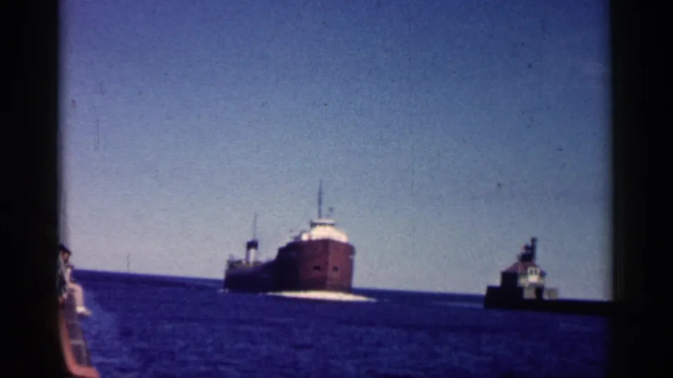 1961 Minnesota home-movie frame with a freighter on the horizon and a small harbor light or structure to the right.