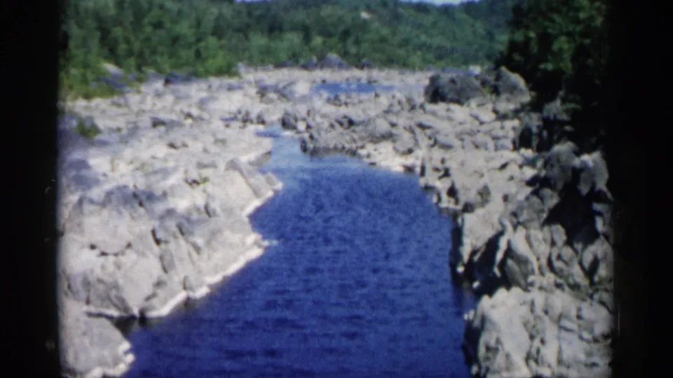 A slightly softer 1961 home-movie frame shows the blue creek curving along pale stone, with green trees beyond the rocky bank.