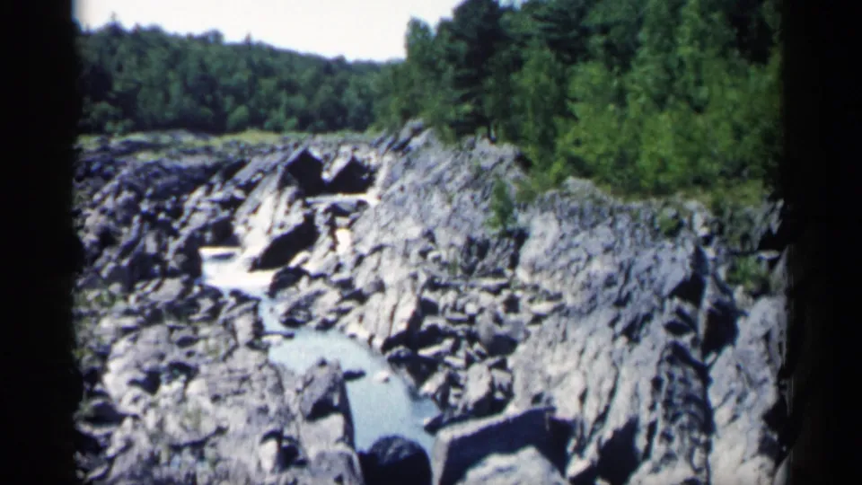 1961 Minnesota 8mm frame showing a rugged stream bend, gray rocks, and dense summer foliage.