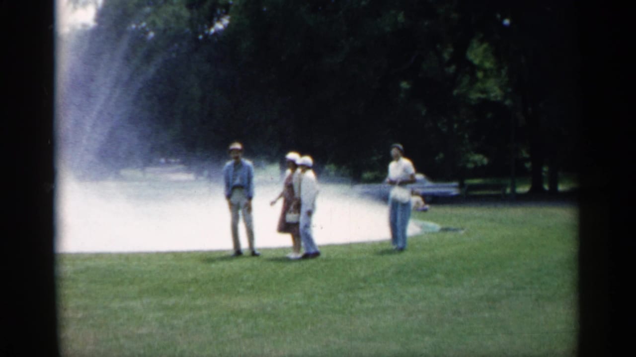 1961 Minnesota 8mm frame of adults in summer clothing standing on grass beside a large park fountain and trees.