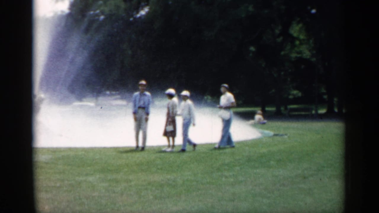 Adults in early-1960s attire move across a Minnesota park lawn with a broad fountain plume behind them.