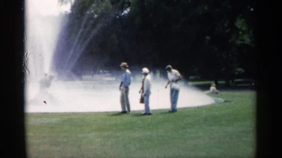 Minnesota home-movie frame from 1961 showing adults standing near a spraying park fountain with trees beyond.