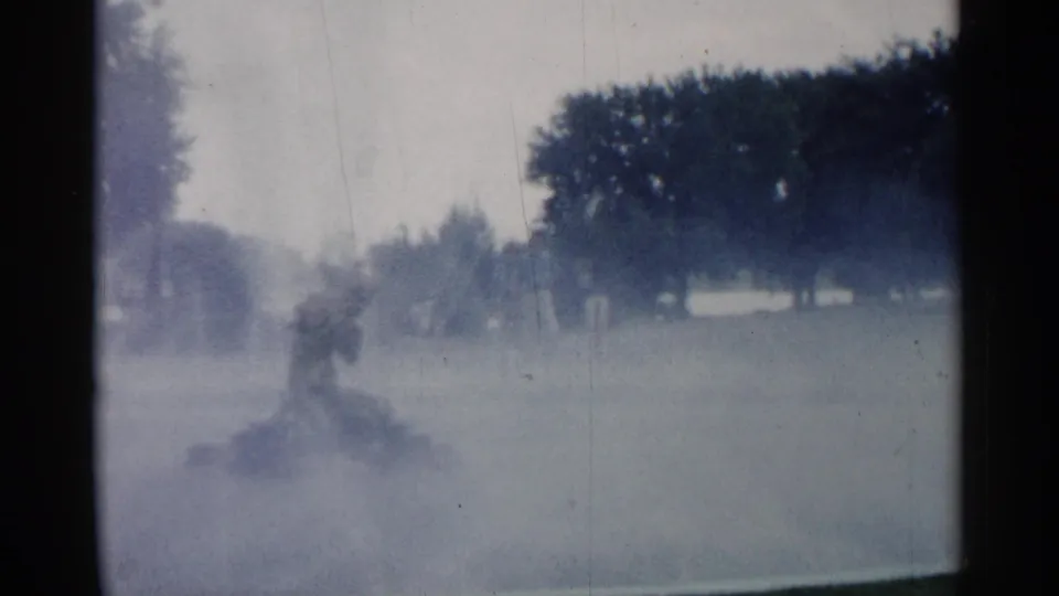 1961 Minnesota 8mm frame of a park lawn where a distant person appears through fog or sprinkler mist near trees.