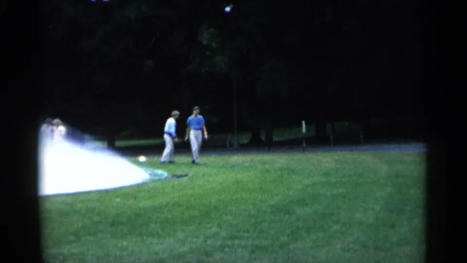 1961 Minnesota home-movie frame showing two adult men walking across a green park lawn beside sprinkler spray.
