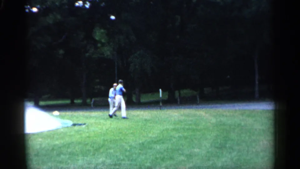 1961 Minnesota 8mm frame with two adult men crossing a park lawn near sprinkler spray and a shaded tree line.
