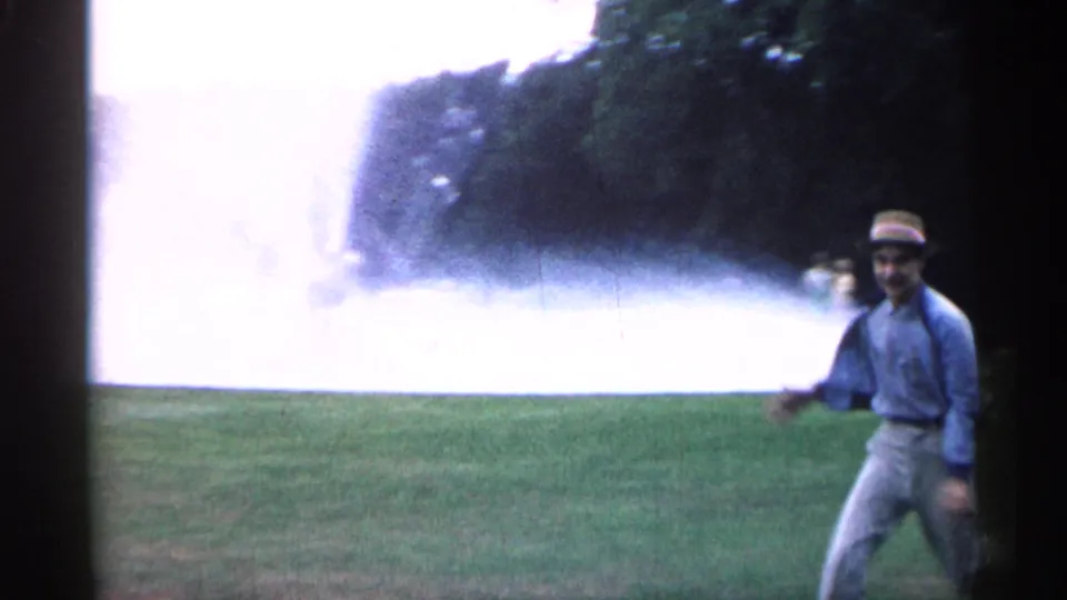 1961 Minnesota frame of a young man in a straw hat and light shirt walking across grass near a large fountain spray.