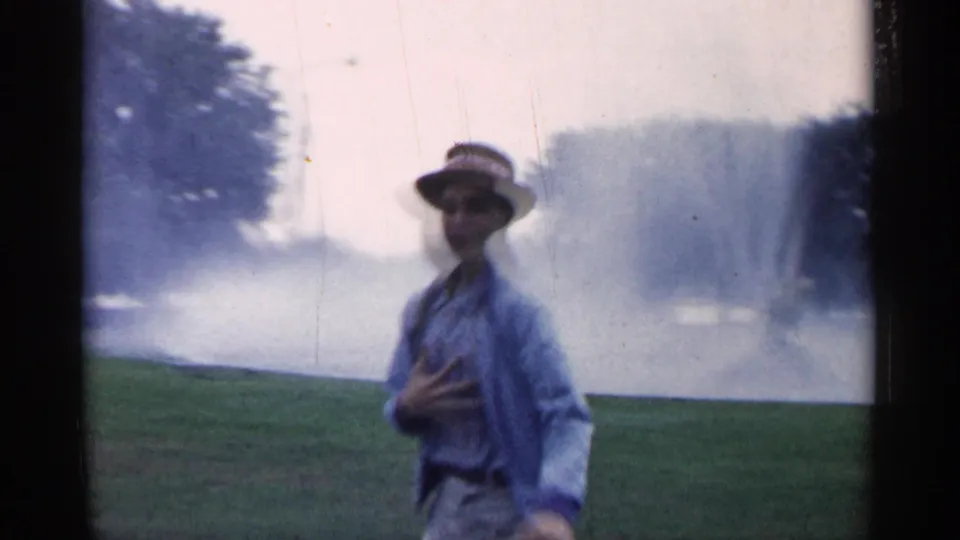 Close 1961 Minnesota home-movie view of the man in a straw hat standing on grass with fountain mist and trees in the background.