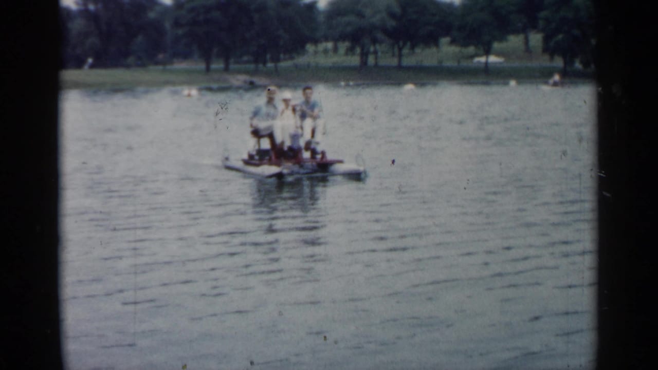 People stand or sit on a small boat on a lake with trees along the far shore.