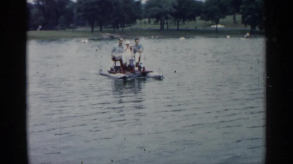 1961 Minnesota home-movie frame showing a small boat with people on a lake, with a tree-lined shoreline in the distance.