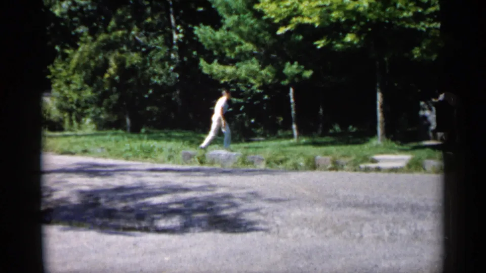 Outdoor Minnesota home-movie frame showing two distant people walking across a paved path bordered by lawn and trees.