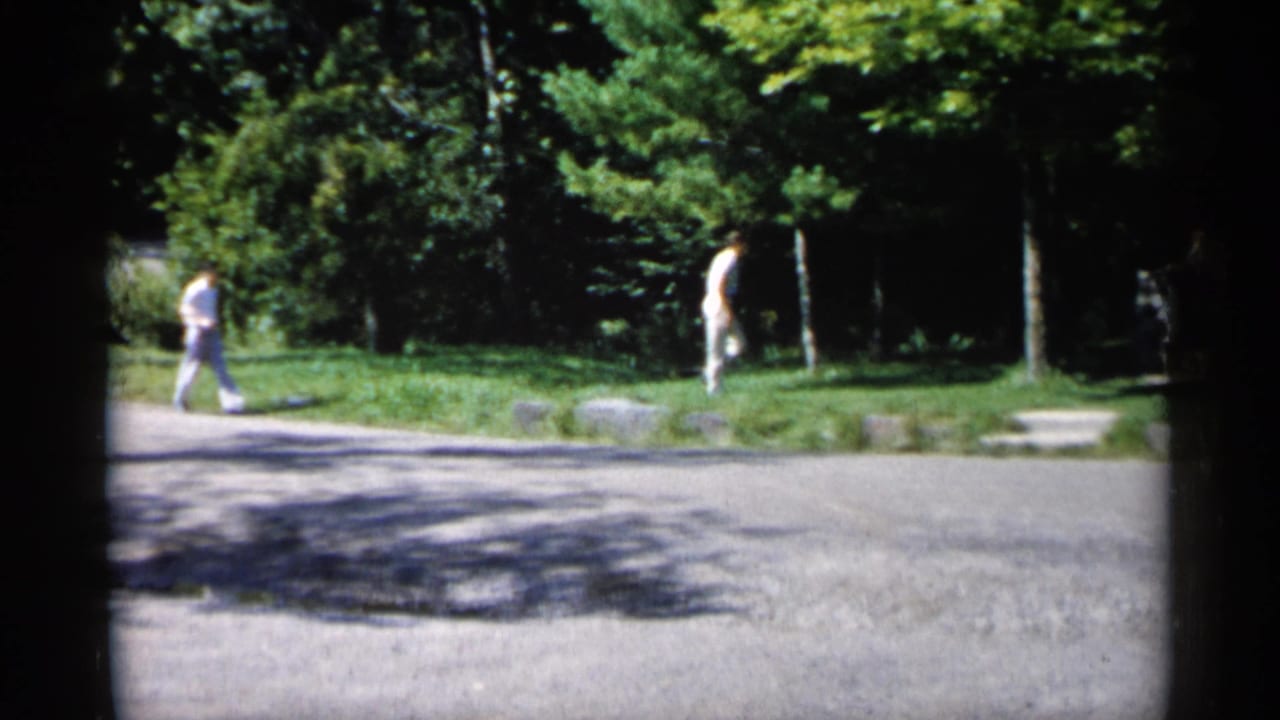 Two figures walk near a paved path and grass in a sunny wooded park.