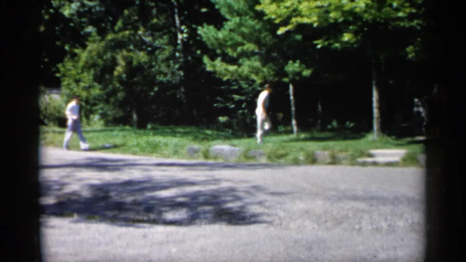 In 1961 Minnesota home-movie footage, two young men appear at a distance near a paved path, grass, and trees in bright sun.