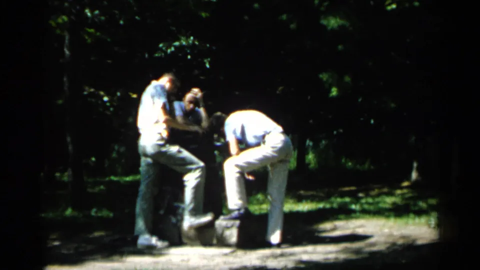 Two young men in light clothing gather near a low stone or block feature in a shaded Minnesota park setting.