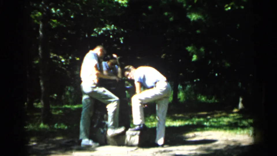 The 1961 park footage shows two young men leaning together near a low stone feature, partly shaded by surrounding trees.
