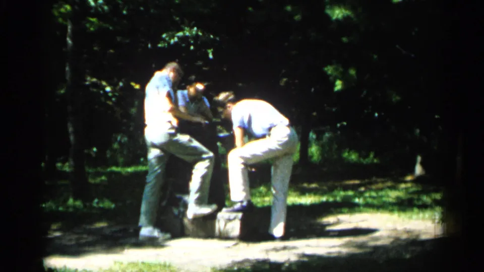 Two young men in light clothing stay clustered near the stone feature in a tree-lined Minnesota park, seen in grainy 8mm color.
