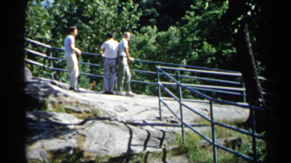 In 1961 Minnesota footage, three adult men in light clothing converse on exposed rock near a blue railing and dense trees.