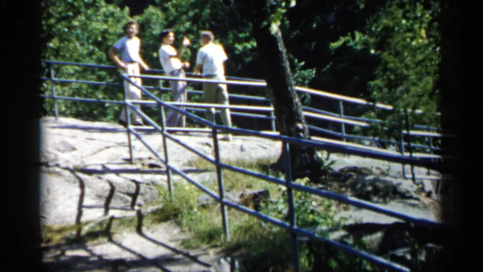 Three adult men appear in conversation near a blue metal railing on a wooded Minnesota rocky outcrop in 1961 home-movie footage.